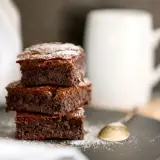 Stos puszystych kostek ciasta brownie z płynną czekoladą, posypanych cukrem pudrem, obok białego kubka i łyżeczki.