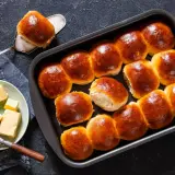 Złociste bułki brioche w blaszce, jedna na łopatce. Obok talerzyk z kostkami masła i nóż. Idealne na śniadanie.