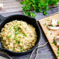 Risotto z grzybami i natką pietruszki w czarnej patelni, obok pokrojone pieczarki na desce.