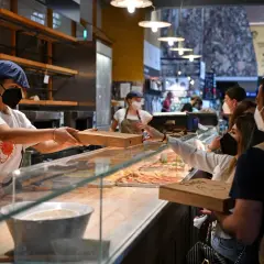 Sprzedawczyni podaje klientce pizzę w pudełku. W tle widać inne pizze i ludzi. Ciekawi Cię, ile kosztuje pizza we Włoszech?