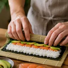 Ręce zwijają nori z ryżem, łososiem i awokado. Obok butelka octu ryżowego do sushi.