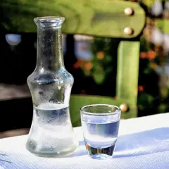 Szklana butelka i kieliszek z uzo na białym obrusie. W tle zielone krzesło i rozmyty ogród.