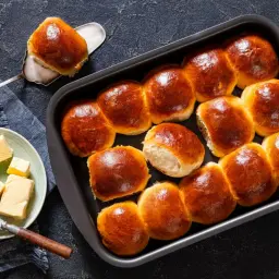 Złociste bułki brioche w blaszce, jedna na łopatce. Obok talerzyk z kostkami masła i nóż. Idealne na śniadanie.