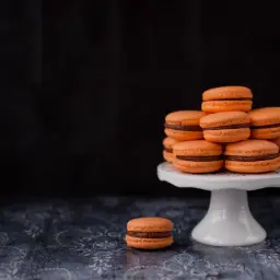 Pomarańczowe makaroniki francuskie z czekoladowym nadzieniem ułożone na białej paterze. Jeden makaronik leży obok.
