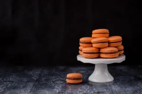 Pomarańczowe makaroniki francuskie z czekoladowym nadzieniem ułożone na białej paterze. Jeden makaronik leży obok.