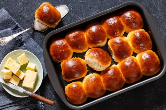 Złociste bułki brioche w blaszce, jedna na łopatce. Obok talerzyk z kostkami masła i nóż. Idealne na śniadanie.