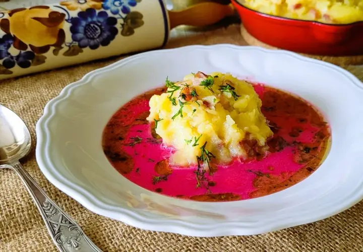 Pyszny barszcz czerwony zabielany, przepis babci. Na talerzu puree ziemniaczane z dodatkiem boczku i koperku, zanurzone w intensywnie różowym barszczu.