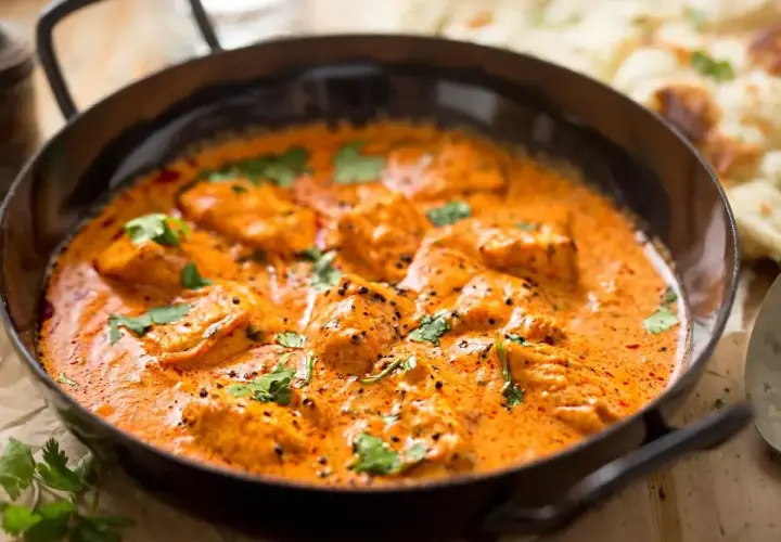 Aksamitny kurczak w sosie butter masala, ozdobiony świeżą kolendrą, podany w czarnej patelni. Obok widać kawałki chleba naan.
