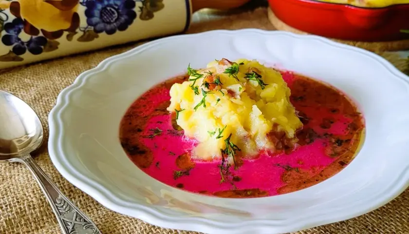 Pyszny barszcz czerwony zabielany, przepis babci. Na talerzu puree ziemniaczane z dodatkiem boczku i koperku, zanurzone w intensywnie różowym barszczu.