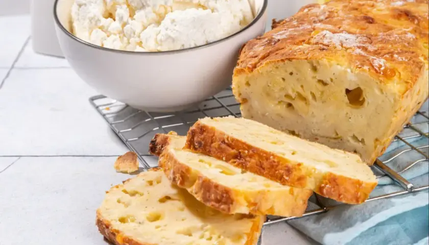 Pyszny chleb z twarogu, świeżo upieczony, z widocznym serem w środku. Obok miska pełna twarogu i butelka oliwy.