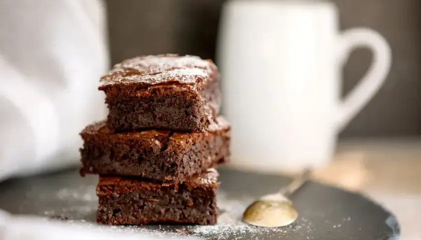 Stos puszystych kostek ciasta brownie z płynną czekoladą, posypanych cukrem pudrem, obok białego kubka i łyżeczki.