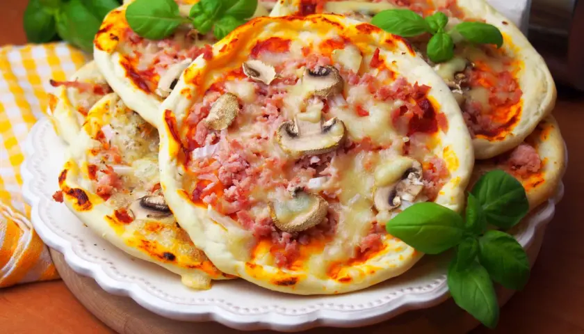 Szybkie mini pizze z szynką i pieczarkami, ozdobione świeżą bazylią, kuszą apetycznym wyglądem.