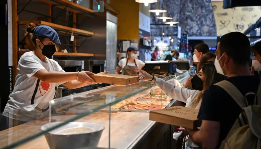 Sprzedawczyni podaje klientce pizzę w pudełku. W tle widać inne pizze i ludzi. Ciekawi Cię, ile kosztuje pizza we Włoszech?