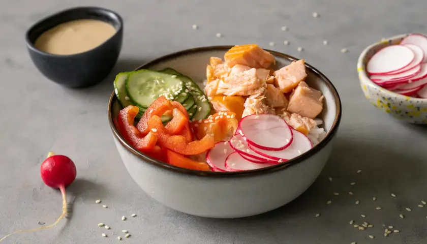 Kolorowa sałatka do sushi z łososiem, ryżem, ogórkiem, papryką i rzodkiewką. Obok miseczka z sosem.