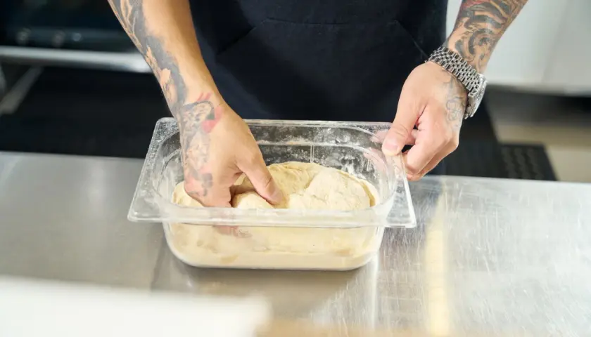 Ręce z tatuażami zagniatają ciasto na pizzę w pojemniku. Czy można mrozic ciasto na pizze? Tak, to dobry sposób na przechowywanie.