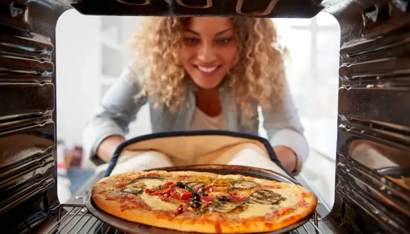 Uśmiechnięta kobieta wyjmuje z piekarnika pyszną pizzę. Widać, na jakim programie piec pizze, bo jest idealnie wypieczona.