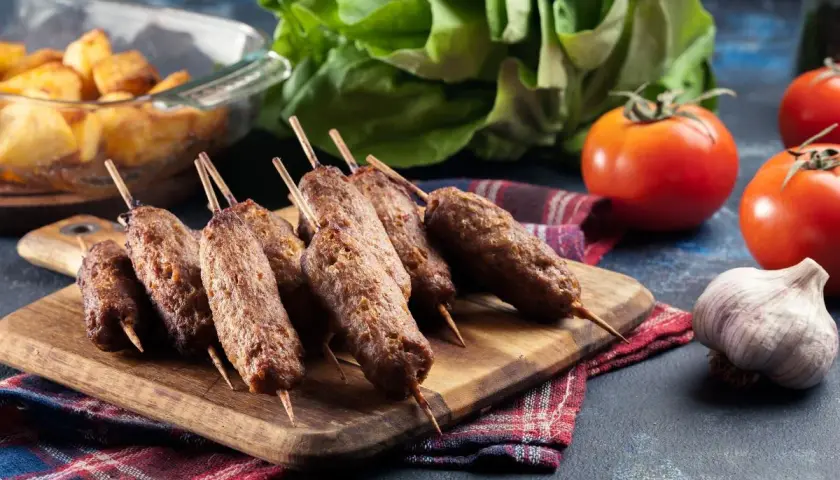 Grillowane ćevapi na drewnianej desce, obok pomidorów, czosnku i pieczonych ziemniaków.