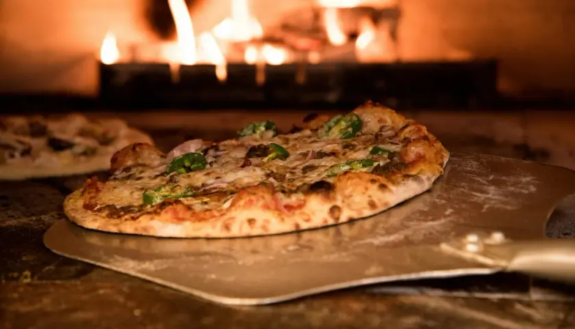 Pizza z pomidorami z puszki, serem i papryczkami jalapeno na łopacie, w tle płomienie pieca.
