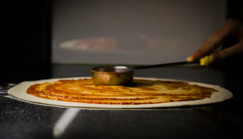 Rozsmarowywanie sosu pomidorowego na cieście pizzy. To kluczowy etap leżakowania ciasta na pizzę, przygotowujący je do dalszych etapów.