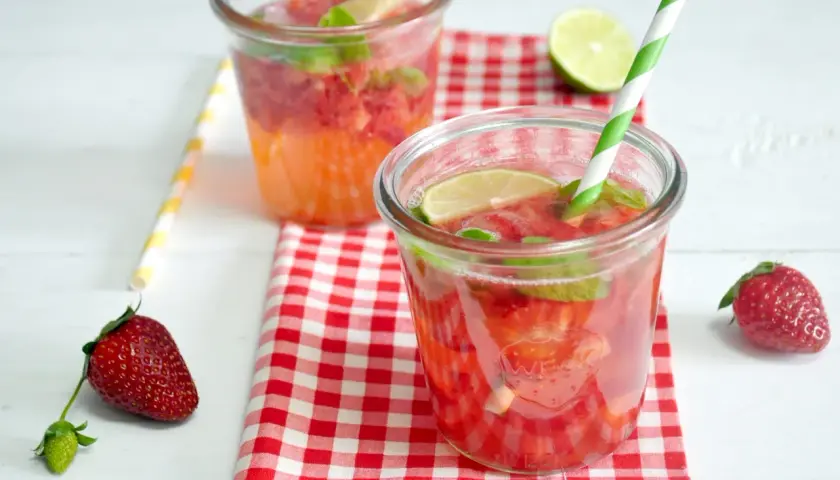 Orzeźwiający drink z truskawkami, miętą i limonką, podany w szklance z zielono-białą słomką. Obok leży świeża truskawka.