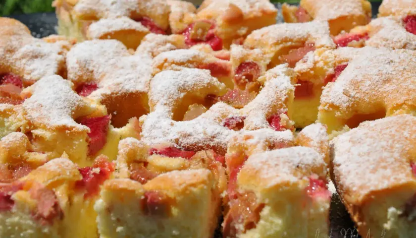 Szybkie ciasto jogurtowe z rabarbarem, posypane cukrem pudrem. Kawałki ciasta z widocznymi różowymi kawałkami rabarbaru.