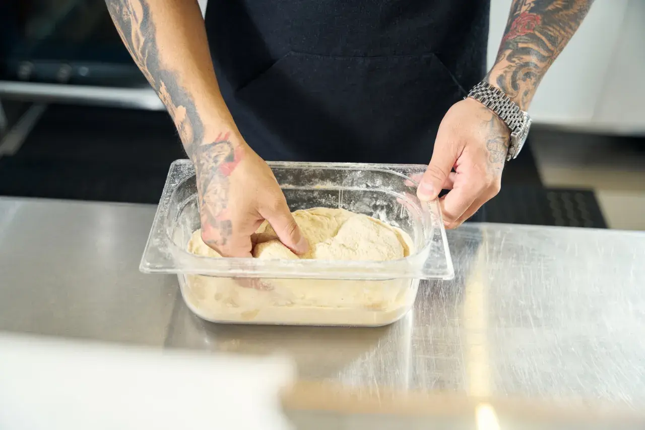 Ręce z tatuażami zagniatają ciasto na pizzę w pojemniku. Czy można mrozic ciasto na pizze? Tak, to dobry sposób na przechowywanie.