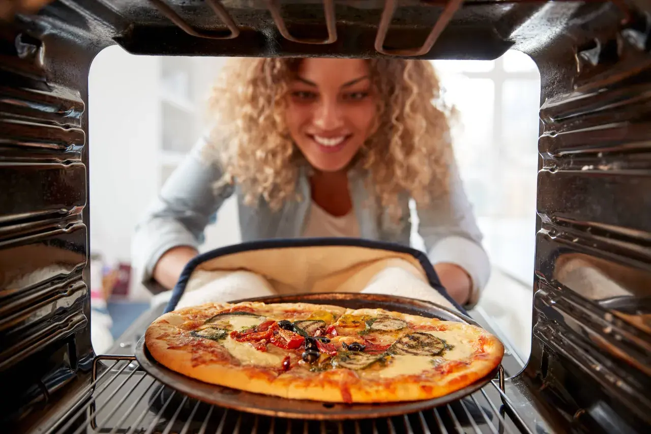 Uśmiechnięta kobieta wyjmuje z piekarnika pyszną pizzę. Widać, na jakim programie piec pizze, bo jest idealnie wypieczona.