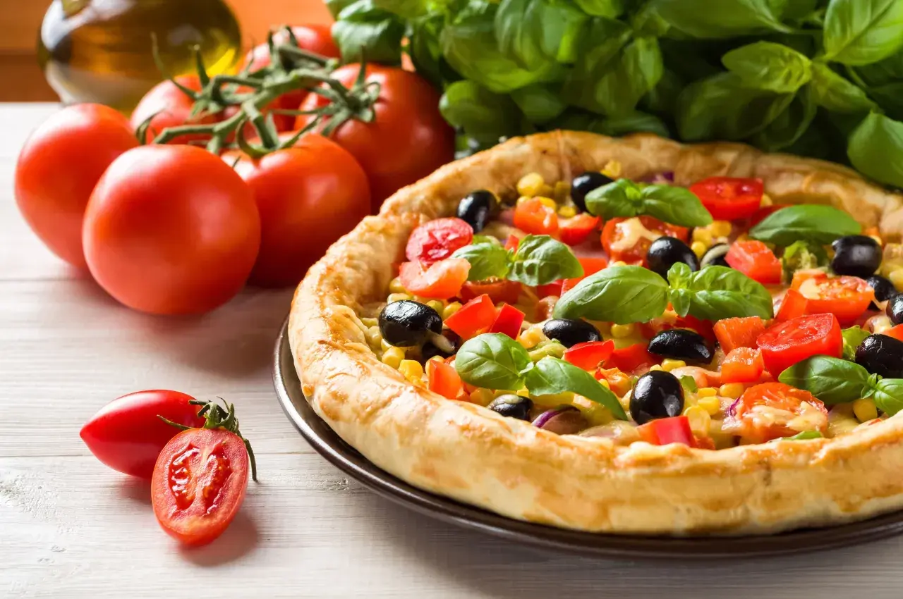 Domowa pizza z pomidorami, oliwkami i kukurydzą, gotowa do pieca. Obok świeże pomidory i bazylia.