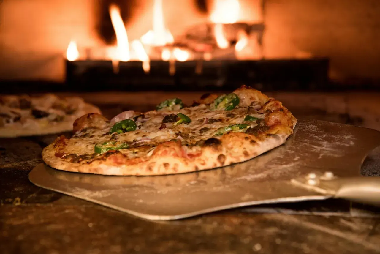 Pizza z pomidorami z puszki, serem i papryczkami jalapeno na łopacie, w tle płomienie pieca.