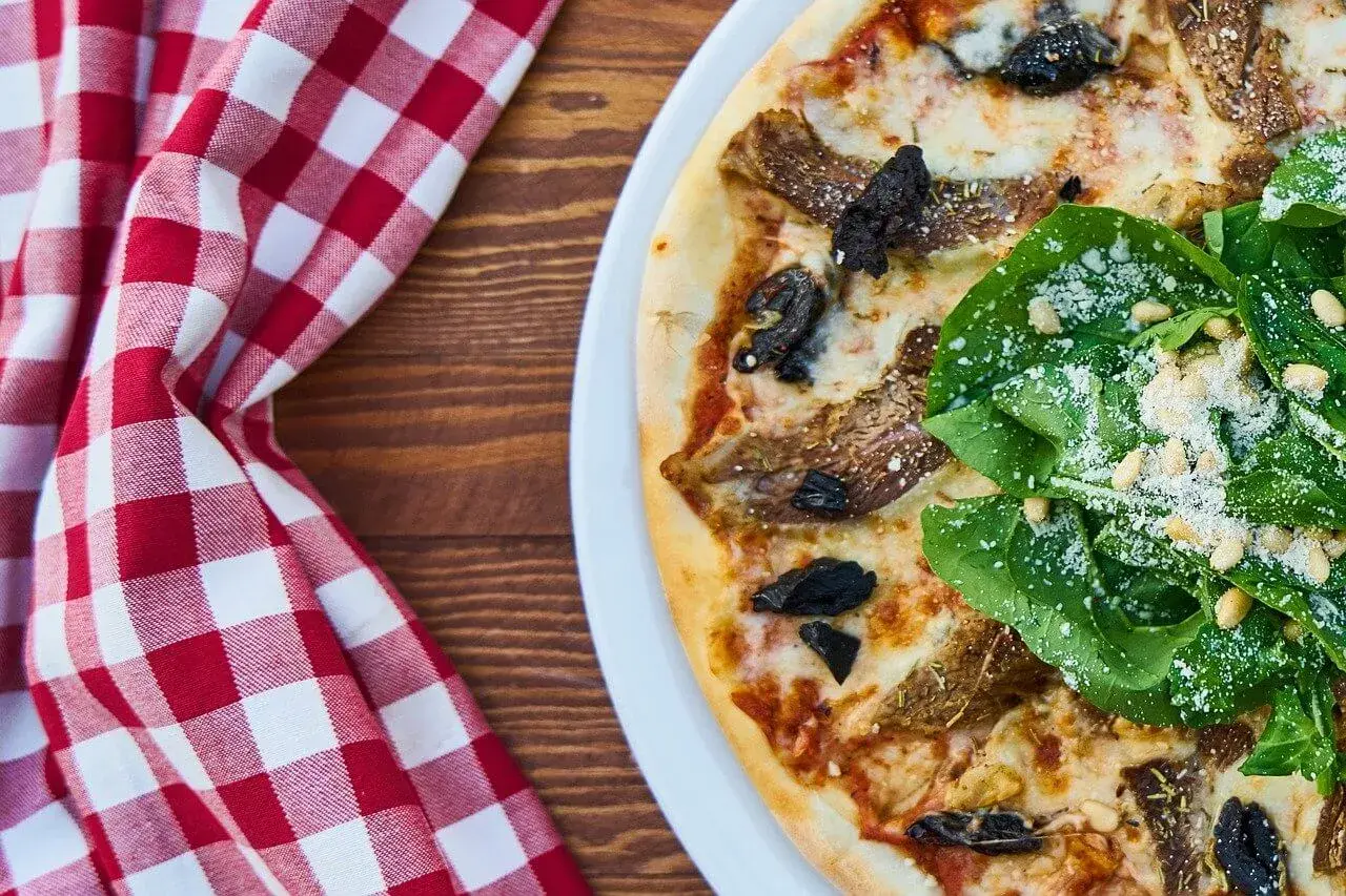 Pizza z rukolą, orzechami pinii i grzybami. Widać kolejność składników na pizzy, od sosu po świeże dodatki.
