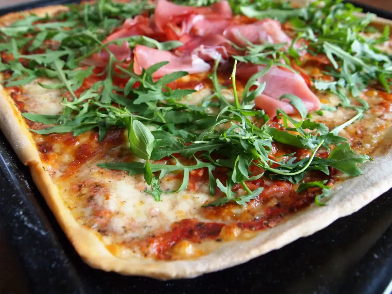 Pizza z rukolą i szynką parmeńską. Widać sos pomidorowy, ser, zielone liście rukoli i różowe plastry szynki. Kolejnosc skladnikow na pizze jest kluczowa dla smaku.